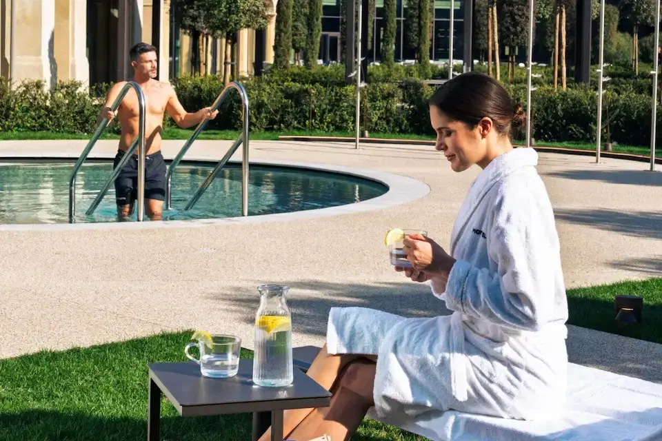 Coppia che si gode un giorno alla spa; la donna si rilassa in accappatoio con un drink, l'uomo nuota in piscina