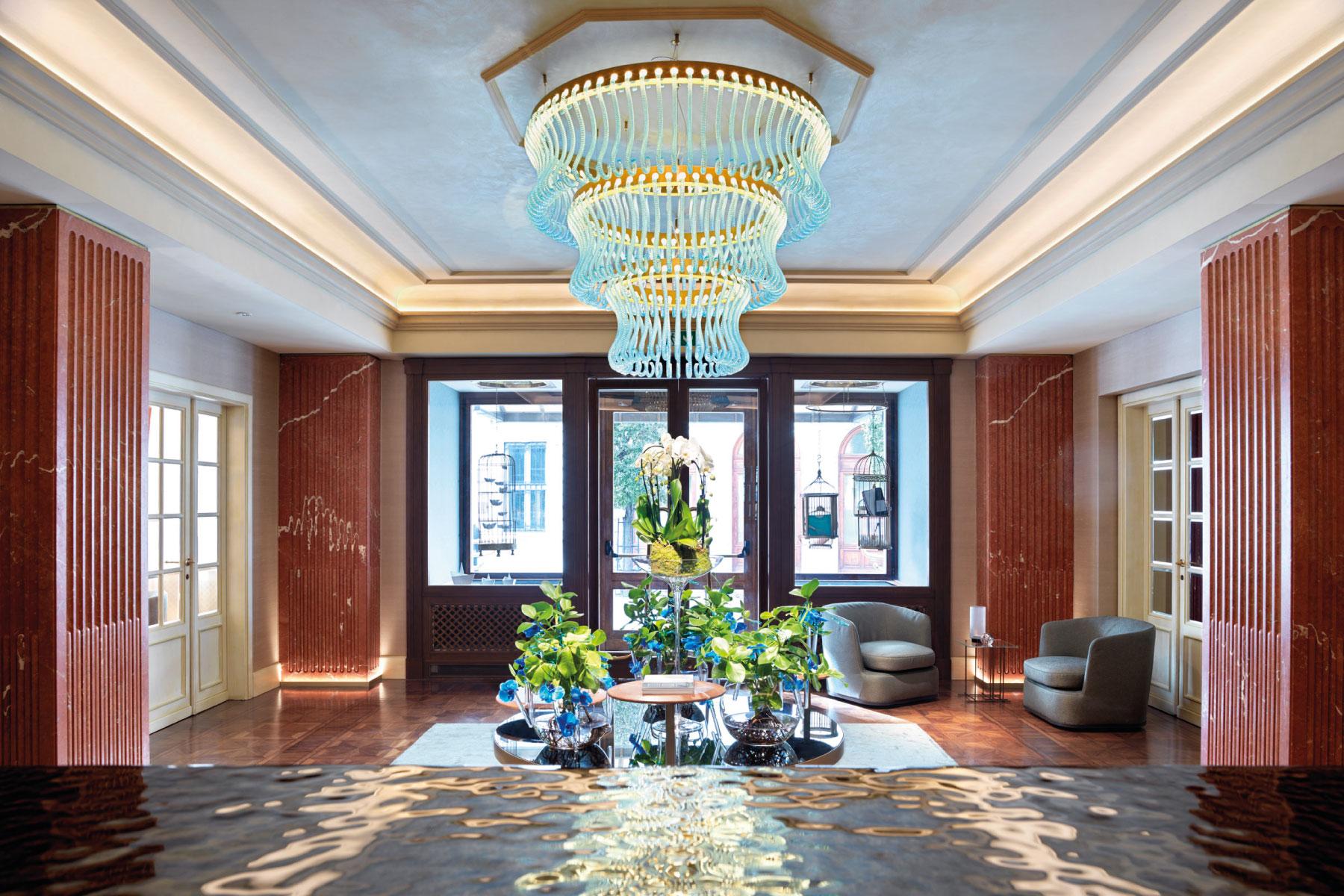 Lussuoso ingresso con lampadario azzurro, tavolino centrale, piante e finestre panoramiche