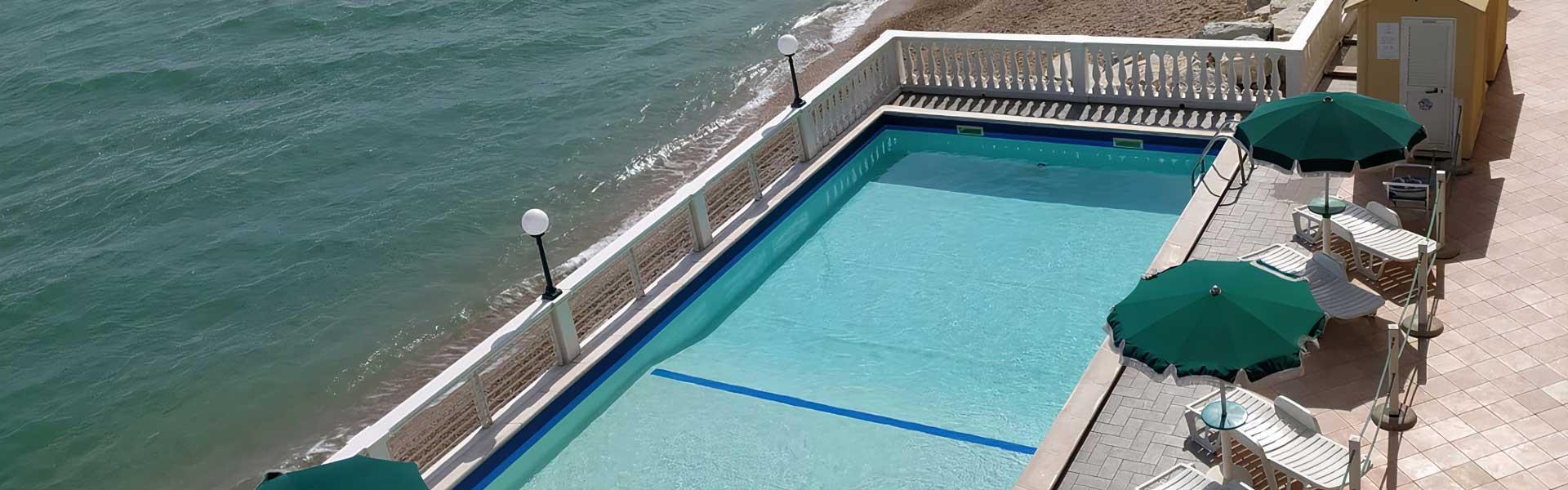 Piscina all'aperto con vista sul mare, ombrelloni verdi e lettini prendisole