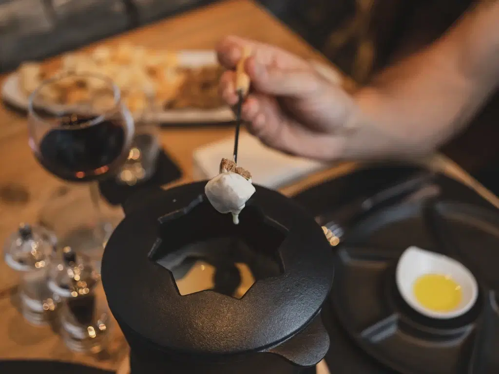 Una persona intinge un pezzo di pane nel formaggio fuso con un bicchiere di vino rosso sul tavolo