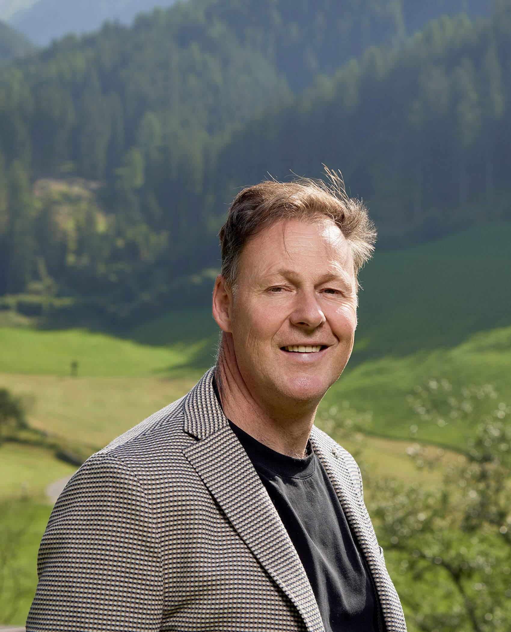 Uomo sorridente in blazer scozzese in piedi all'aperto con colline verdi e alberi sullo sfondo