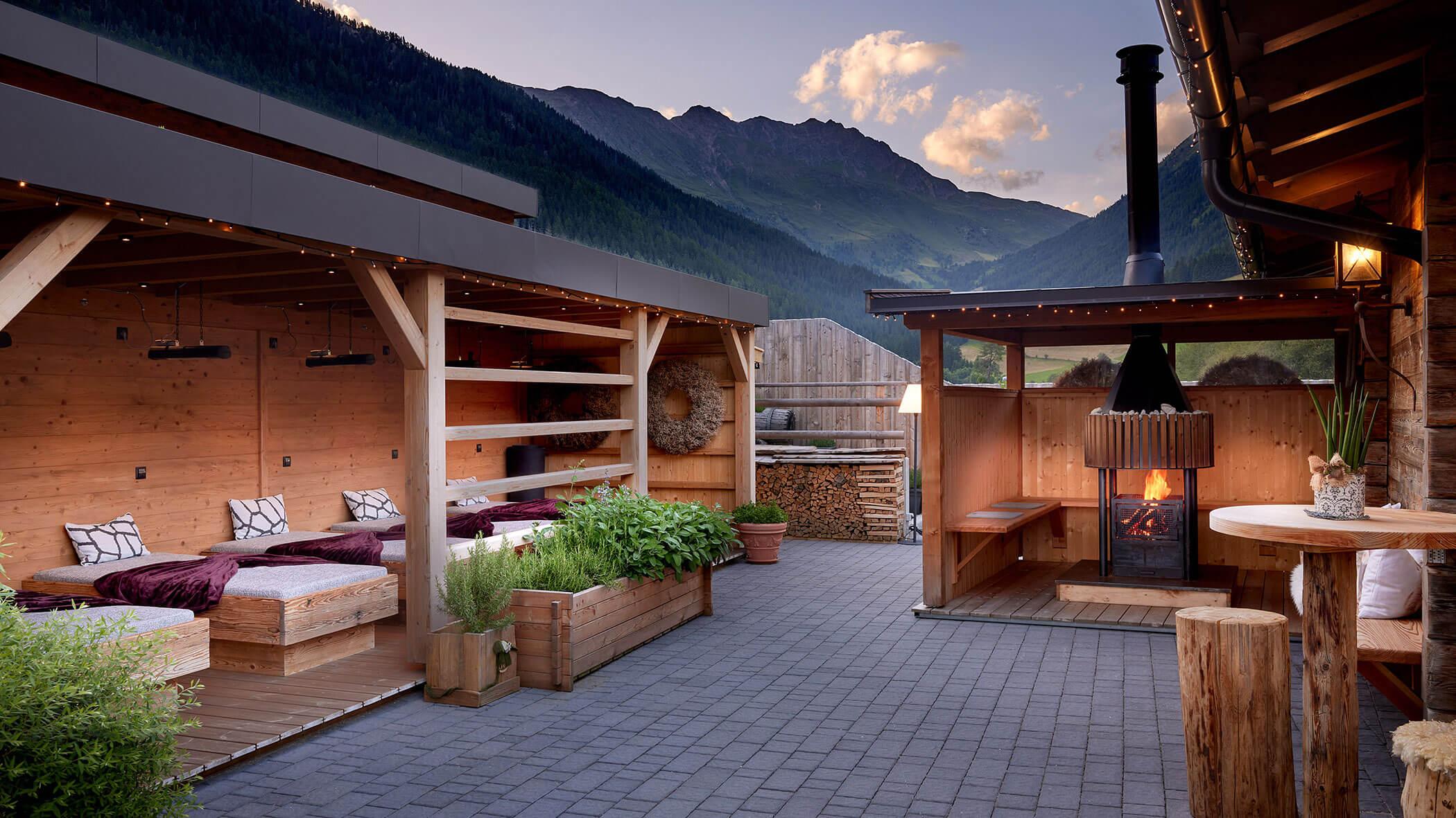 Terrazza esterna accogliente con sedute in legno, camino e vista sulle montagne al tramonto