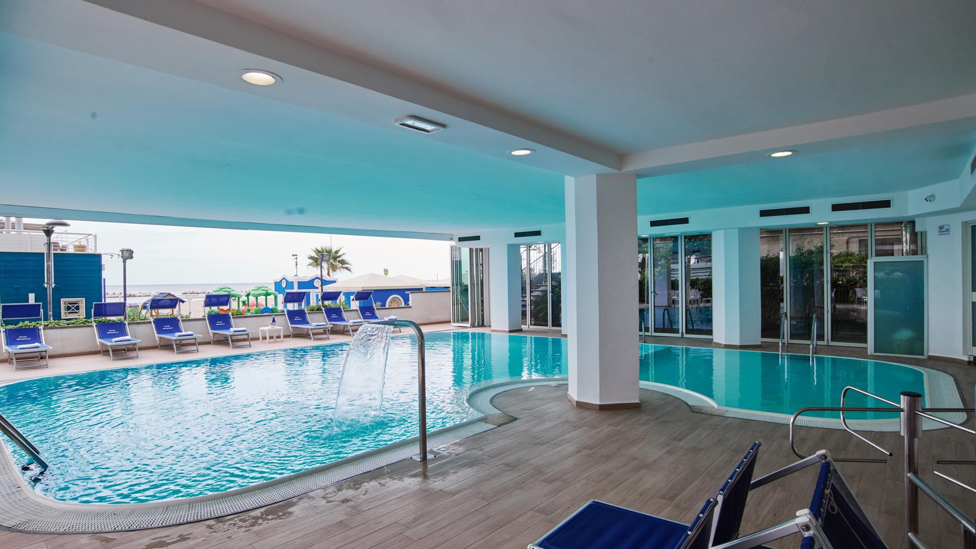 Piscina coperta con vista mare, lettini blu e cascata d'acqua